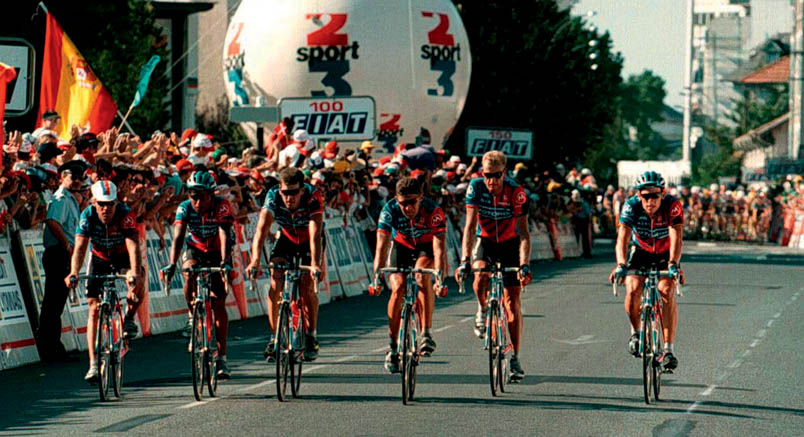 On the background, the peloton waits to let the Motorola team pass as a group in Pau. From left to right: Steve Bauer, Alvaro Mejia, Frankie Andreu, Andrea Peron, Stephen Swart, and Lance Armstrong. It is Peron who has received the instruction 'keep riding' from Casartelli's wife in a phone call. Casartelli's roommate may cross the finish line one meter ahead of the group and thus have the 'stage win' credited to Casartelli.