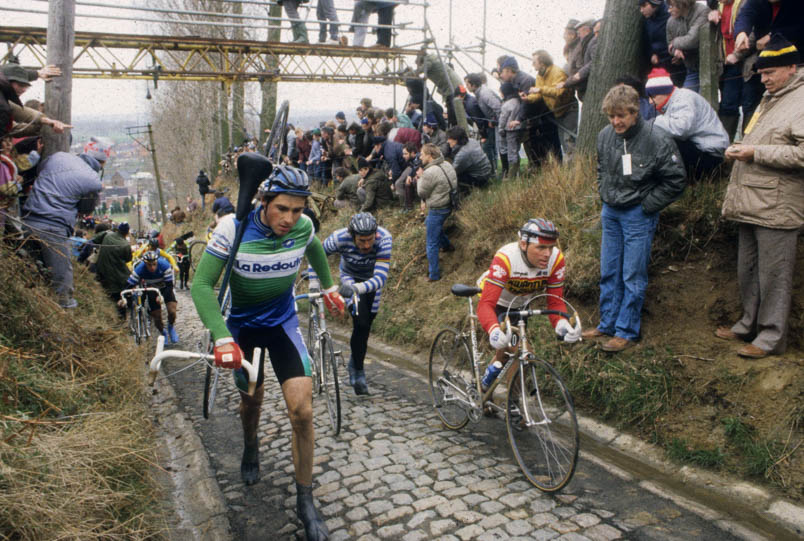 1984 in de Ronde van Vlaanderen. Parool-verslaggever Harrie ten Asbroek (witte kaart om de nek) heeft een plekje op de Koppenberg gevonden om Hennie Kuiper lopend naar boven te zien komen. Links heeft Alain Bondue de fiets op de nek. Achter Hennie kluunt Urs Freuler