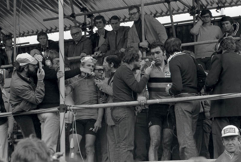 Vreugde en ontgoocheling op de perstribune in Yvoir, 1975. Wereldkampioen Hennie Kuiper wordt geïnterviewd door Theo Koomen (links) en Jean Nelissen (rechts). Links van Koomen probeert Hans Prakke een foto van Kuiper te maken. Naast Kuiper – met bril – NOS-regisseur Martijn Lindenberg. Een luttele meter verder druipt de droefheid Eddy Merckx van het gelaat, wanneer hij de vragen van Fred De Bruyne op zich krijgt afgevuurd.