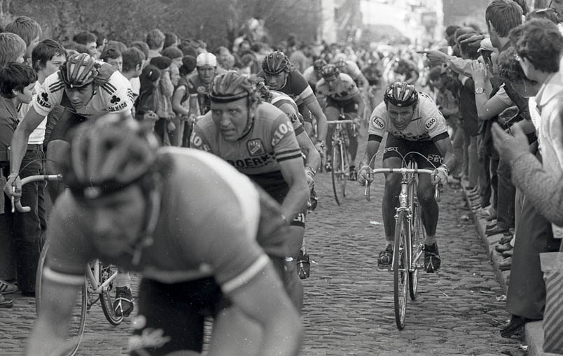 In the 1979 Fleche Wallonne, a tough race is being ridden. Bernard Hinault is almost done. Far left, Fons De Wolf is pushing hard. To the right of De Wolf, Walter Godefroot fixes his gaze on a point in the distance. Just behind Godefroot, Henk Lubberding rides in his national champion jersey. On the right, Hennie Kuiper tries to avoid getting caught up in the struggle.