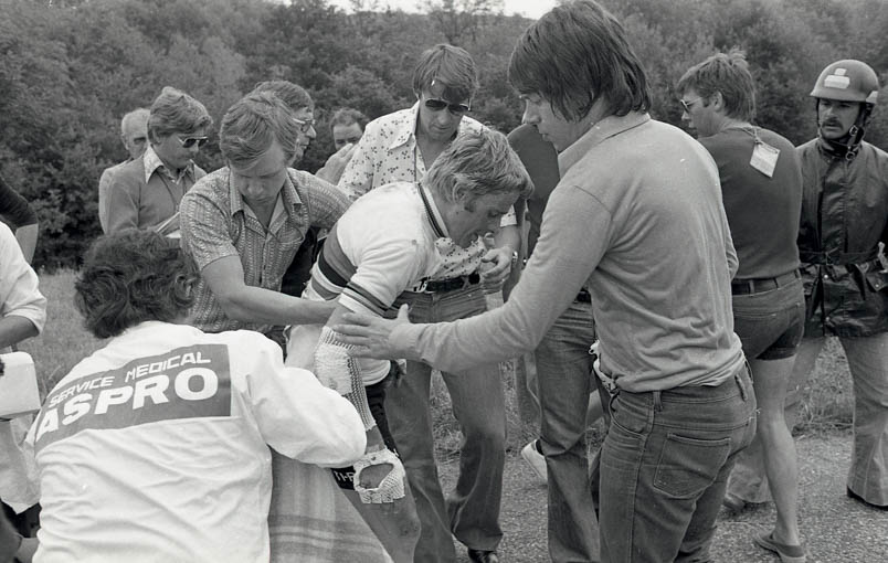 Tourarts Gérard Porte slaat ter ondersteuning beide armen om Hennie Kuiper om hem op de been te helpen. Ook Peter Post (met zonnebril) biedt hulp. Journalisten komen polshoogte nemen: links achter Kuiper Leo van de Ruit van het ANP, daarnaast Theo Koomen en helemaal rechts (met de accreditatie op zijn rug) winnaar van Olympisch brons in Tokio en NOS-chauffeur Gerard Koel