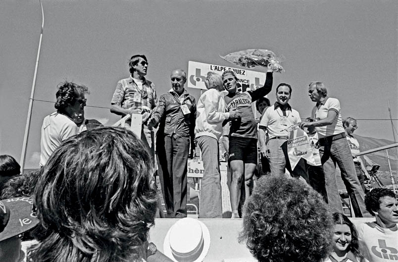 Het podium voor de huldiging van Hennie Kuiper op l’Alpe d’Huez is nog lang niet wat het tegenwoordig is. Toch is het een plek om te zien en gezien te worden. Dit keer valt de eer te beurt aan de Belgische zangeres Annie Cordy om Kuiper te zoenen. Helemaal links op het podium staat – met zonnebril – ook ploegleider Peter Post trots te zijn