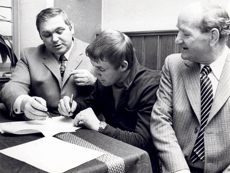 On 19 December 1972, Hennie Kuiper signs his first professional contract. He will ride for Ha-Ro, the team of Robert Kahl, who is pointing to Hennie on the left in the photo where he should sign. Brother Oude Keizer, Hennie's first coach, looks approvingly at where his guidance has led.