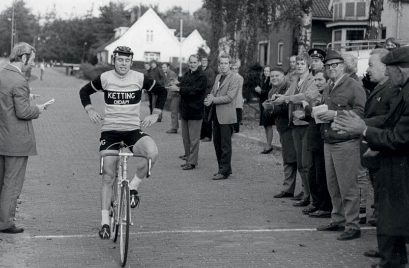 Zichtbaar tevreden passeert Hennie Kuiper de streep bij de clubkampioenschappen in Oldenzaal. Links jurylid Ben Roesink, rechts van Hennie in een zwarte duffelse jas OWC-voorzitter Herman Nijhuis met naast hem (applaudisserend) Willy Wissink